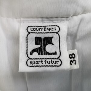 Puede incluir: Primer plano de una etiqueta blanca con la marca "courr&egrave;ges" y el texto "sport futur" en negro. La etiqueta tambi&eacute;n presenta un logotipo negro y el n&uacute;mero "38". La etiqueta est&aacute; adherida a una tela blanca.