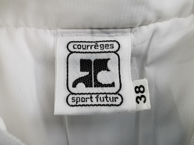 Puede incluir: Primer plano de una etiqueta blanca con la marca "courr&egrave;ges" y el texto "sport futur" en negro. La etiqueta tambi&eacute;n presenta un logotipo negro y el n&uacute;mero "38". La etiqueta est&aacute; adherida a una tela blanca.