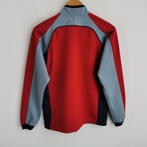 Op de afbeelding: Een rode en grijze sportjas met een zwarte kraag en accenten. De jas heeft een ritssluiting over de hele lengte en lange mouwen. De achterkant van de jas is zichtbaar en toont het colorblock-ontwerp.