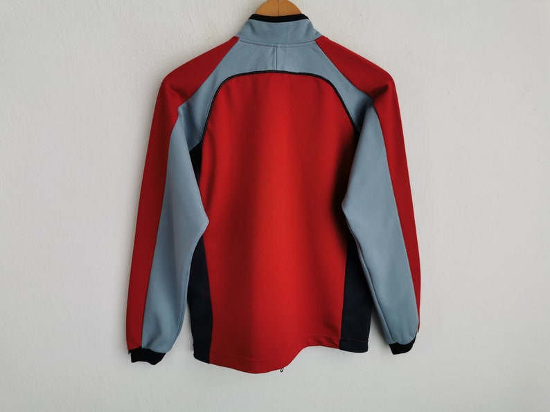 Op de afbeelding: Een rode en grijze sportjas met een zwarte kraag en accenten. De jas heeft een ritssluiting over de hele lengte en lange mouwen. De achterkant van de jas is zichtbaar en toont het colorblock-ontwerp.