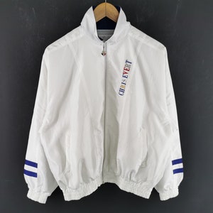 Puede incluir: Chaqueta blanca con cremallera y el texto "CHRIS EVERT" en azul, rojo y amarillo en el pecho izquierdo. La chaqueta tiene rayas azules en las mangas y un cuello azul marino. La chaqueta está colgada en una percha de madera.