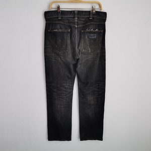 May include: Dark gray denim jeans with a distressed look, hanging on a wooden hanger. The jeans feature a classic five-pocket design, a Wrangler logo on the back pocket, and red stitching details.