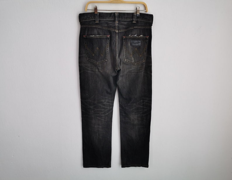 May include: Dark gray denim jeans with a distressed look, hanging on a wooden hanger. The jeans feature a classic five-pocket design, a Wrangler logo on the back pocket, and red stitching details.