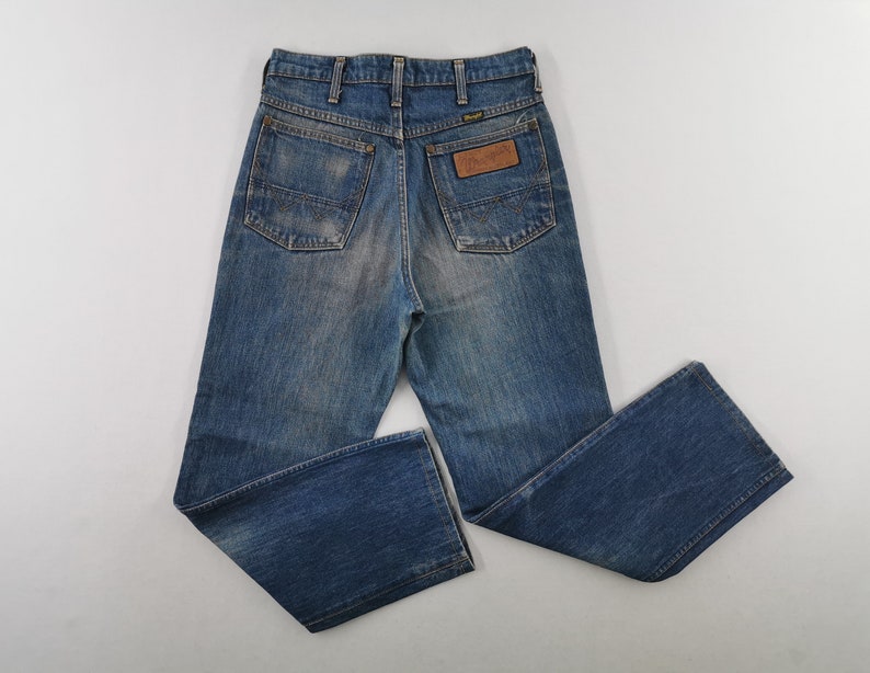 K&ouml;nnte beinhalten: Blaue Jeans aus Denim mit verwaschener Optik. Die Jeans haben ein klassisches F&uuml;nf-Taschen-Design, einen braunen Lederaufn&auml;her mit dem "Wrangler"-Logo und einen geraden Beinverlauf. Die Jeans liegen flach auf einer wei&szlig;en Oberfl&auml;che.