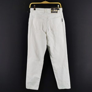 May include: A pair of white denim pants hanging on a wooden hanger against a black background. The pants have a classic five-pocket design and a straight leg. A tag is visible on the back.