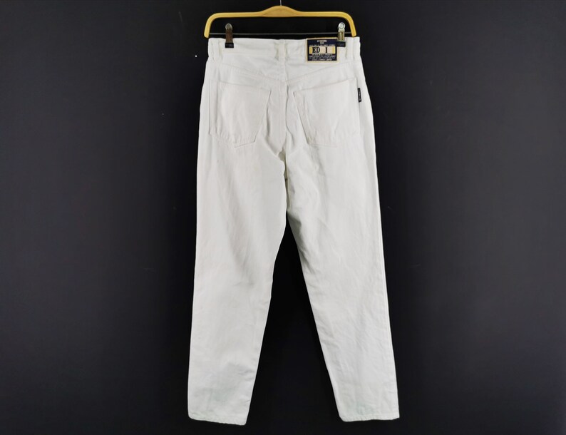 May include: A pair of white denim pants hanging on a wooden hanger against a black background. The pants have a classic five-pocket design and a straight leg. A tag is visible on the back.