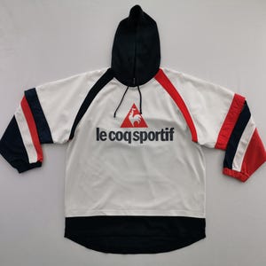 K&ouml;nnte beinhalten: Wei&szlig;er Kapuzenpullover mit schwarzen und roten Akzenten. Auf der Jacke steht der Text "le coq sportif" in Schwarz, mit einem roten Hahn-Logo. Die &Auml;rmel haben rote, wei&szlig;e und marineblaue Streifen. Die Kapuze ist schwarz.