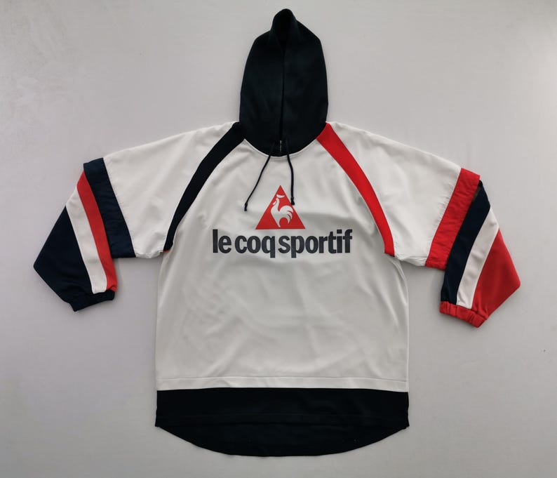 K&ouml;nnte beinhalten: Wei&szlig;er Kapuzenpullover mit schwarzen und roten Akzenten. Auf der Jacke steht der Text "le coq sportif" in Schwarz, mit einem roten Hahn-Logo. Die &Auml;rmel haben rote, wei&szlig;e und marineblaue Streifen. Die Kapuze ist schwarz.