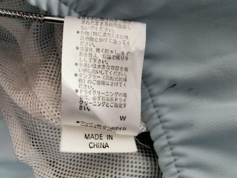 May include: A close-up of a clothing tag with Japanese text, a white mesh fabric, and a light blue fabric. The tag reads "MADE IN CHINA".