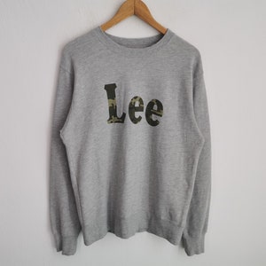 May include: A heather grey crewneck sweatshirt featuring the word Lee printed across the chest in a dark green, brown, and black camouflage pattern. The garment has long sleeves with ribbed cuffs and a ribbed hem, displayed on a wooden hanger.