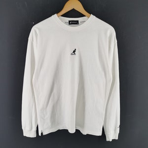 Peut inclure: T-shirt blanc &agrave; manches longues et col rond. Il pr&eacute;sente un petit logo kangourou noir sur la poitrine et un petit logo sur la manche. Le t-shirt est suspendu &agrave; un cintre en bois sur fond sombre.