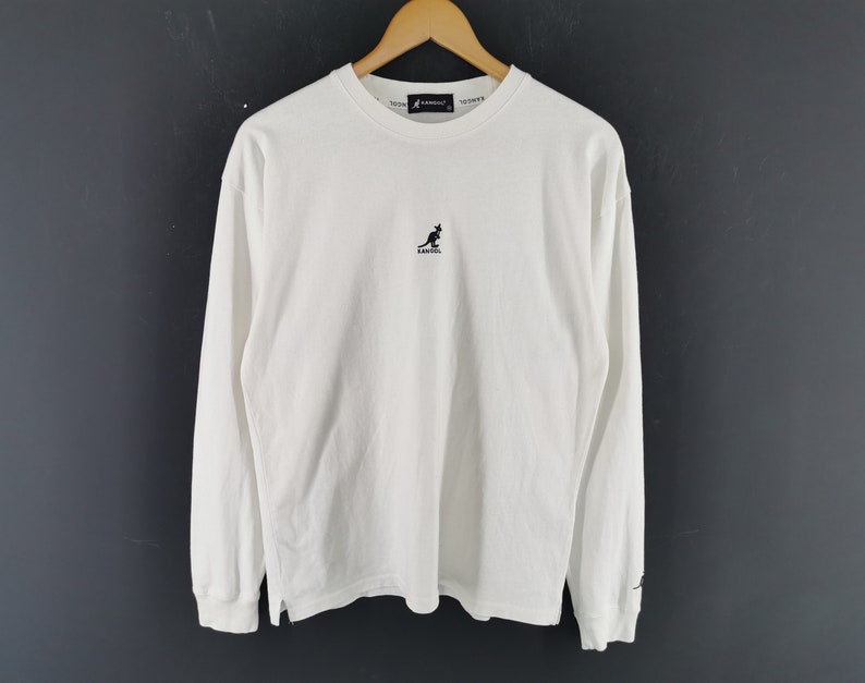 Peut inclure: T-shirt blanc &agrave; manches longues et col rond. Il pr&eacute;sente un petit logo kangourou noir sur la poitrine et un petit logo sur la manche. Le t-shirt est suspendu &agrave; un cintre en bois sur fond sombre.