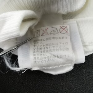 May include: Close-up of a white fabric label with Japanese text and care symbols. The label is attached to a white garment. The text includes instructions for washing and ironing. A silver tool is pointing at the label.