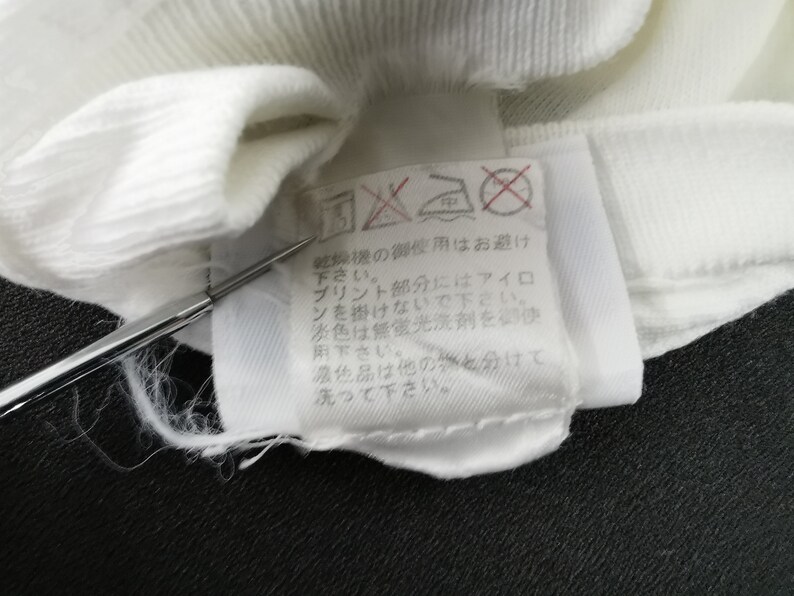 May include: Close-up of a white fabric label with Japanese text and care symbols. The label is attached to a white garment. The text includes instructions for washing and ironing. A silver tool is pointing at the label.