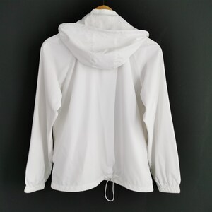 Puede incluir: Chaqueta blanca con capucha y mangas largas, vista desde atr&aacute;s. La chaqueta tiene un cord&oacute;n en la cintura y cierre de cremallera. Cuelga de una percha de madera sobre un fondo oscuro.
