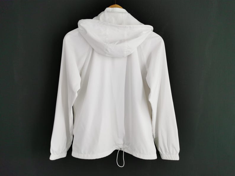 Puede incluir: Chaqueta blanca con capucha y mangas largas, vista desde atr&aacute;s. La chaqueta tiene un cord&oacute;n en la cintura y cierre de cremallera. Cuelga de una percha de madera sobre un fondo oscuro.