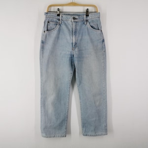 Può includere: Jeans di denim azzurro appesi a una gruccia di legno. I jeans hanno un classico design a cinque tasche, una chiusura con bottone e cerniera e una vestibilità dritta. Il denim ha un lavaggio sbiadito.