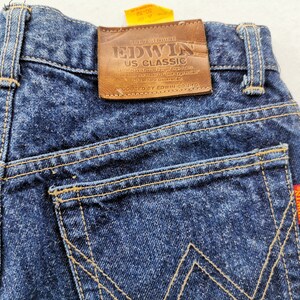 May include: Close-up of the back of dark blue denim jeans. The jeans feature a brown leather patch with "EDWIN US CLASSIC" branding. The back pockets have a distinctive stitching pattern, and a red tag with "EDWIN US CLASSIC" is visible.