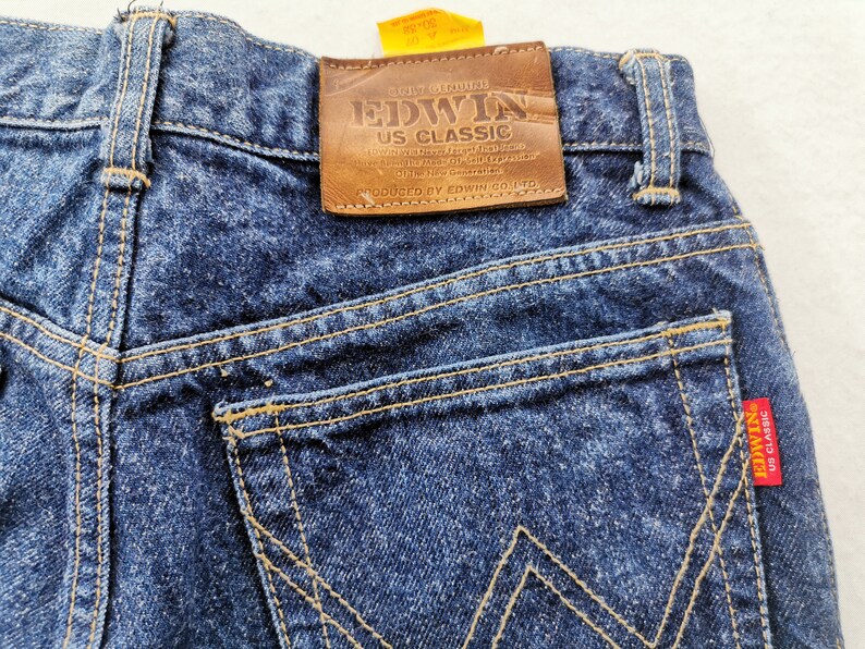 May include: Close-up of the back of dark blue denim jeans. The jeans feature a brown leather patch with "EDWIN US CLASSIC" branding. The back pockets have a distinctive stitching pattern, and a red tag with "EDWIN US CLASSIC" is visible.