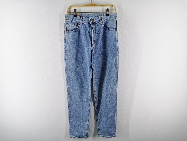 May include: Light blue denim jeans hanging on a wooden hanger. The jeans have a classic five-pocket design, a button and zipper closure, and a straight leg fit. The jeans are made of a durable denim material.