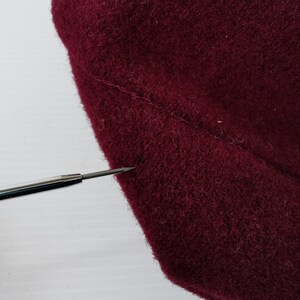 May include: Close-up of a section of burgundy fabric, possibly wool or felt, showing a seam. A silver tool with a pointed tip is positioned near the fabric, highlighting the texture and color. The fabric appears soft and slightly textured.