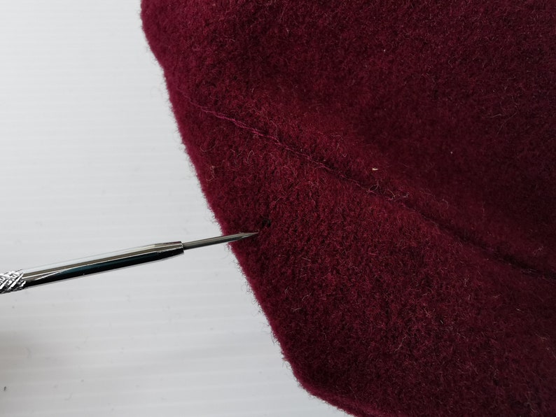 May include: Close-up of a section of burgundy fabric, possibly wool or felt, showing a seam. A silver tool with a pointed tip is positioned near the fabric, highlighting the texture and color. The fabric appears soft and slightly textured.