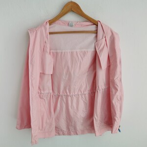 May include: A light pink windbreaker jacket featuring a full-length front zipper, a drawstring waist, and long sleeves. The interior of the jacket is lined with a white mesh fabric, and the garment is displayed on a wooden clothes hanger.