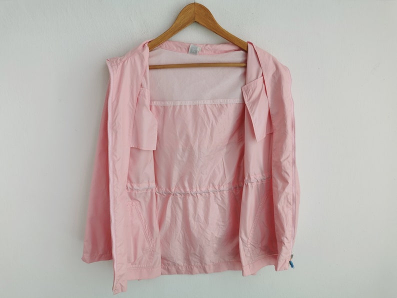 May include: A light pink windbreaker jacket featuring a full-length front zipper, a drawstring waist, and long sleeves. The interior of the jacket is lined with a white mesh fabric, and the garment is displayed on a wooden clothes hanger.