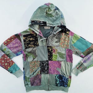 Puede incluir: Una sudadera con capucha de patchwork con cremallera y capucha. La chaqueta presenta una variedad de cuadrados de tela coloridos en tonos de azul, morado, verde y negro. Las mangas tienen bolsillos y puños de canalé. La marca "BULANA" es visible en la etiqueta.