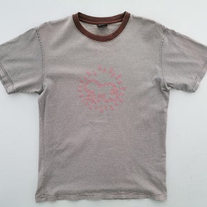 Pode incluir: T-shirt de manga curta &agrave;s riscas cinza e castanho. Tem gola castanha e um desenho gr&aacute;fico rosa na frente, possivelmente um sol ou um cora&ccedil;&atilde;o estilizado. A t-shirt &eacute; feita de material macio.