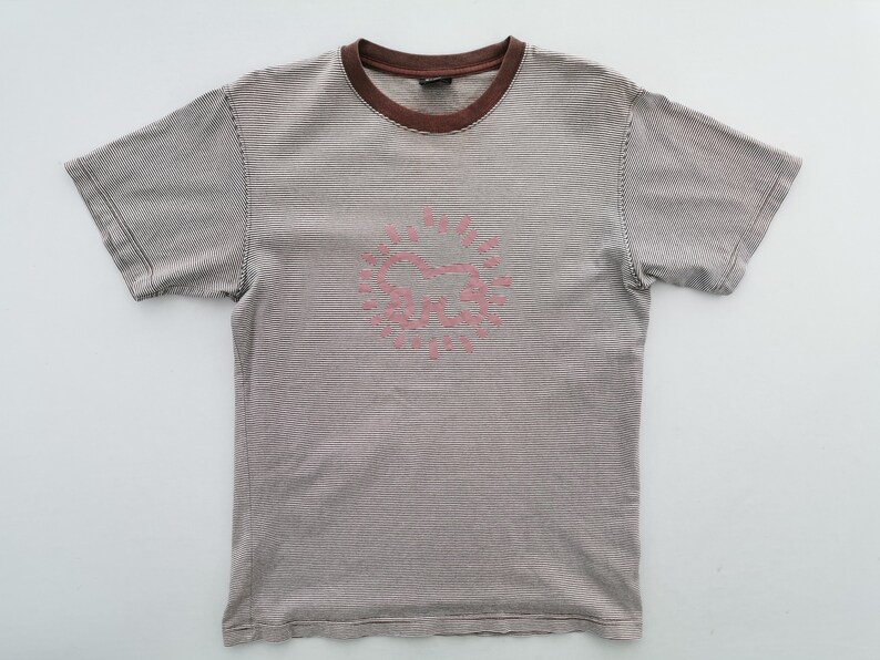 Pode incluir: T-shirt de manga curta &agrave;s riscas cinza e castanho. Tem gola castanha e um desenho gr&aacute;fico rosa na frente, possivelmente um sol ou um cora&ccedil;&atilde;o estilizado. A t-shirt &eacute; feita de material macio.