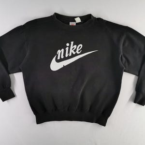 K&ouml;nnte beinhalten: Schwarzes Sweatshirt mit Rundhalsausschnitt und wei&szlig;em Nike-Logo und Swoosh-Design auf der Brust. Das lang&auml;rmlige Oberteil hat gerippte B&uuml;ndchen und einen gerippten Saum. Das Nike-Logo ist in Schreibschrift.