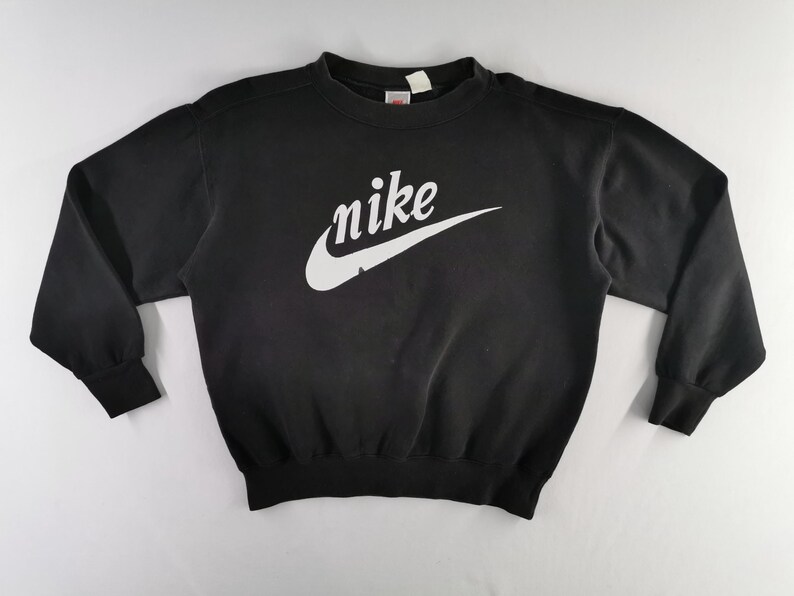 K&ouml;nnte beinhalten: Schwarzes Sweatshirt mit Rundhalsausschnitt und wei&szlig;em Nike-Logo und Swoosh-Design auf der Brust. Das lang&auml;rmlige Oberteil hat gerippte B&uuml;ndchen und einen gerippten Saum. Das Nike-Logo ist in Schreibschrift.
