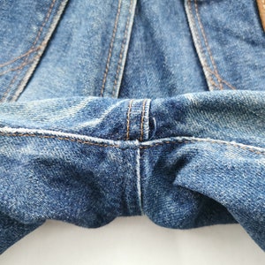 Peut inclure: Gros plan sur un jean en denim bleu, mettant en valeur les coutures et la texture du tissu. L'image met en &eacute;vidence les d&eacute;tails du v&ecirc;tement, y compris les coutures et l'aspect us&eacute; du denim. L'&eacute;tiquette Wrangler est visible.