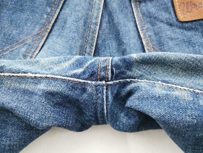 Peut inclure: Gros plan sur un jean en denim bleu, mettant en valeur les coutures et la texture du tissu. L'image met en &eacute;vidence les d&eacute;tails du v&ecirc;tement, y compris les coutures et l'aspect us&eacute; du denim. L'&eacute;tiquette Wrangler est visible.