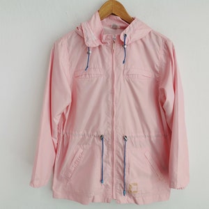 May include: A light pink hooded windbreaker jacket featuring a full-length front zipper, long sleeves, and two front pockets. The garment includes blue drawstring cords at the hood and waist, with a small rectangular patch on the lower front.