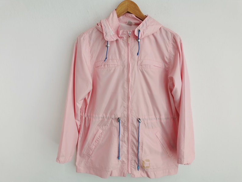 May include: A light pink hooded windbreaker jacket featuring a full-length front zipper, long sleeves, and two front pockets. The garment includes blue drawstring cords at the hood and waist, with a small rectangular patch on the lower front.