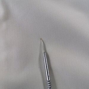 Puede incluir: Primer plano de una tela gris claro con superficie texturizada. Una herramienta plateada en forma de aguja est&aacute; colocada en la esquina inferior derecha, con una peque&ntilde;a mancha marr&oacute;n cerca de la punta.