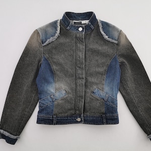 Puede incluir: Chaqueta vaquera en tonos grises y azules. La chaqueta presenta un cuello alzado, cierre de botones en la parte delantera y dos bolsillos delanteros. Los hombros y los puños tienen un detalle deshilachado. La chaqueta es una mezcla de lavados vaqueros.