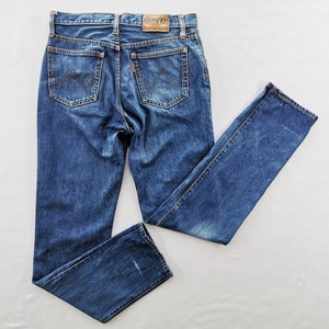 Può includere: Jeans in denim blu con un classico design a cinque tasche. I jeans presentano un lavaggio sbiadito e una toppa in pelle con il nome del marchio. Le tasche posteriori hanno un motivo di cucitura distintivo. I jeans sono stesi su una superficie bianca.