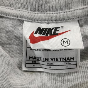 May include: Close-up of a gray t-shirt collar with a Nike label. The label features the Nike logo in black and red, the size is M, and the text "MADE IN VIETNAM".