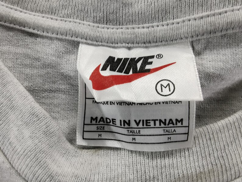 May include: Close-up of a gray t-shirt collar with a Nike label. The label features the Nike logo in black and red, the size is M, and the text "MADE IN VIETNAM".