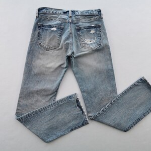 May include: Light blue denim jeans with distressed details, including rips on the back pockets and knees. The jeans have a classic five-pocket design and a faded wash, giving them a worn-in appearance. The jeans are laid flat on a white surface.