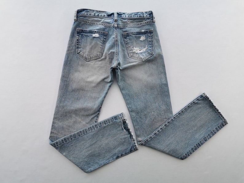 May include: Light blue denim jeans with distressed details, including rips on the back pockets and knees. The jeans have a classic five-pocket design and a faded wash, giving them a worn-in appearance. The jeans are laid flat on a white surface.
