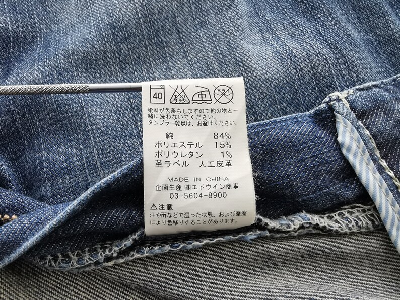 May include: Close-up of blue denim fabric with a sewn-in label. The label provides washing instructions and fabric composition: 84% cotton, 15% polyester, and 1% polyurethane. The label also indicates the product was made in China.
