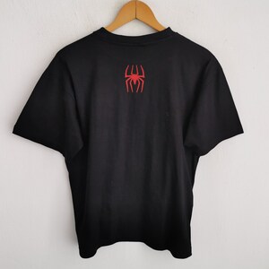 Pode incluir: Camiseta preta com um emblema de aranha vermelha nas costas. A camiseta est&aacute; pendurada em um cabide de madeira contra uma parede branca. O emblema &eacute; um design de aranha estilizado.