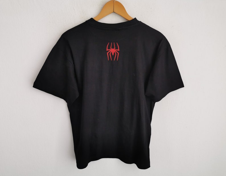 Pode incluir: Camiseta preta com um emblema de aranha vermelha nas costas. A camiseta est&aacute; pendurada em um cabide de madeira contra uma parede branca. O emblema &eacute; um design de aranha estilizado.