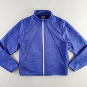 May include: A blue zip-up jacket with a textured, grid-like pattern. The jacket has a stand-up collar and a white zipper. Dark blue stitching accents the front and sleeves. The jacket is a solid blue color.