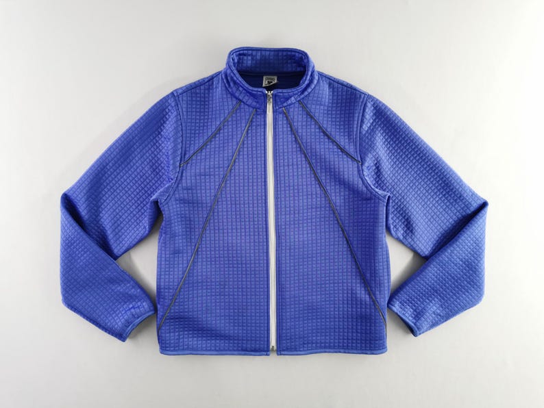 May include: A blue zip-up jacket with a textured, grid-like pattern. The jacket has a stand-up collar and a white zipper. Dark blue stitching accents the front and sleeves. The jacket is a solid blue color.