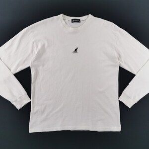 Peut inclure: Un t-shirt blanc cass&eacute; &agrave; manches longues et col rond. Le t-shirt pr&eacute;sente un petit logo kangourou noir centr&eacute; sur la poitrine et le nom de la marque imprim&eacute; sur le col. Les manches sont longues et les poignets sont c&ocirc;tel&eacute;s.